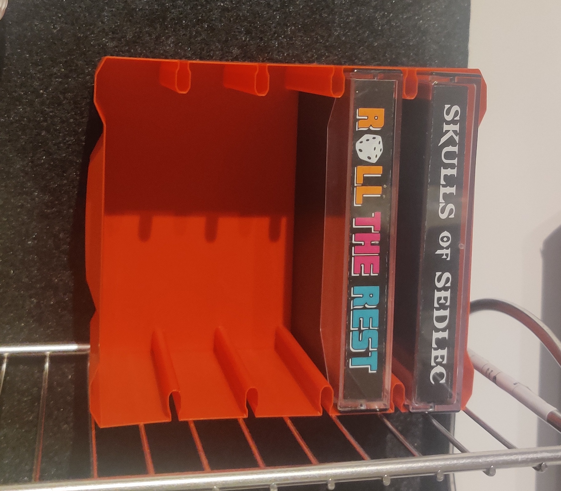 A bright red 3D printed container with grooves that holds 5 C cassette cases. Two of the slots are filled with cassette cases, one reads Roll the Rest and another reads Skulls of Sedlec on their spines.