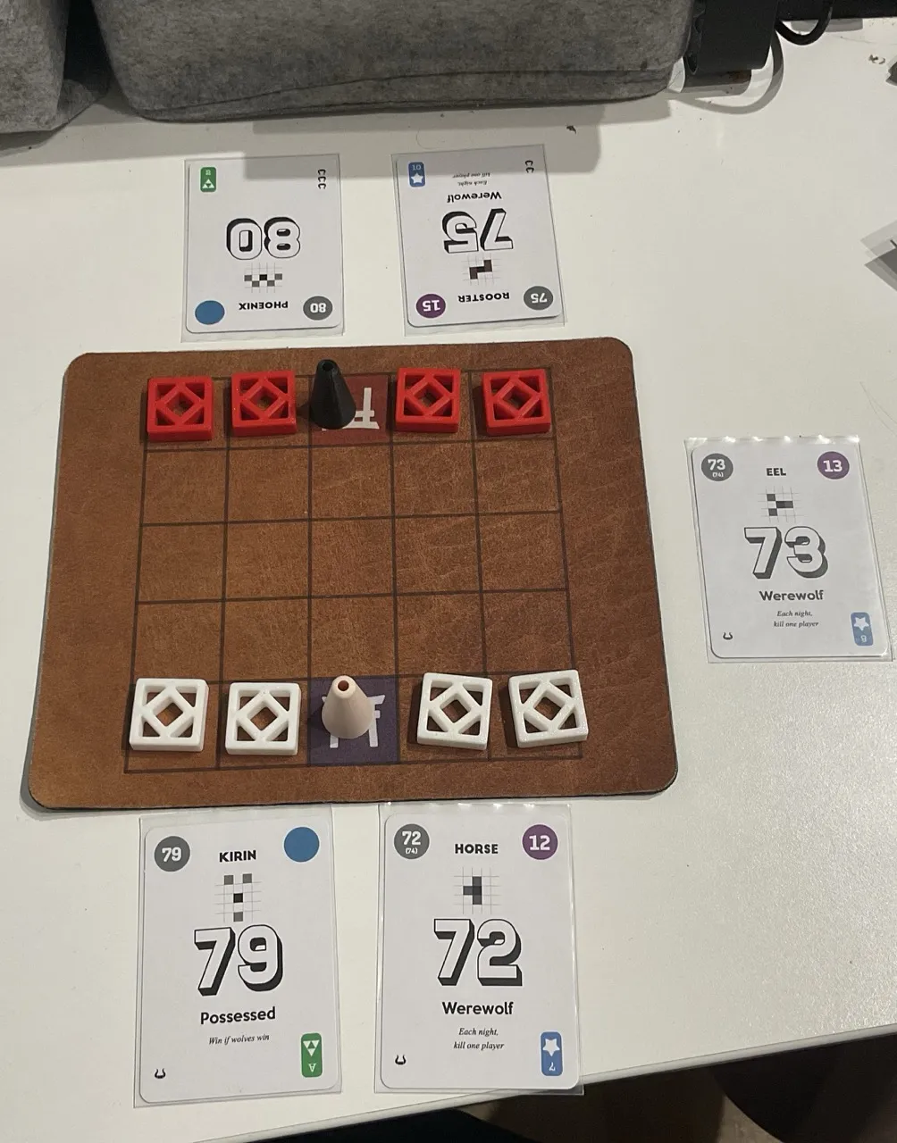 A starting position of Onitama but with 3D printed white and red components and a brown mousepad with a 5 by 5 grid printed on it. The onmyo pawns are represented by black and beige 3D printed cones.