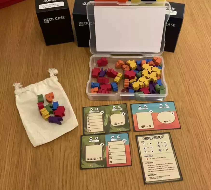 A small photo box full of colourful animal meeples, three poker card size custom player mats and a reference card for Draftosaurus