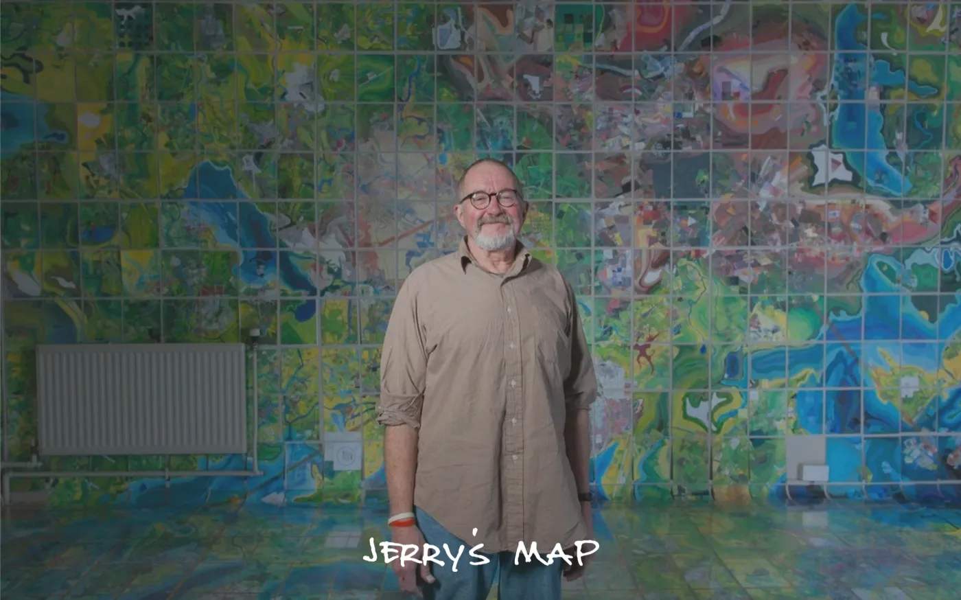 A man standing in front of a large wall covered with a map built by tiles he has painted. The map is very green and blue and red.