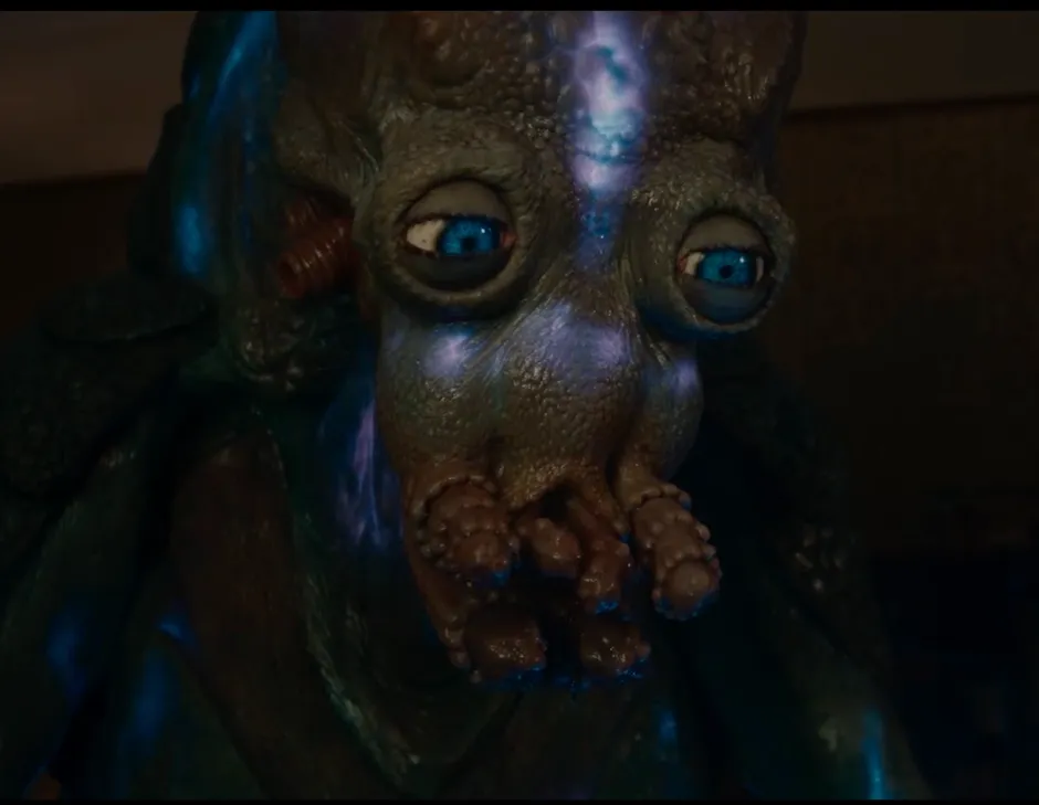 A dark green movie alien with bright blue eyes and tentacle-like mouth. It looks kind and compassionate.