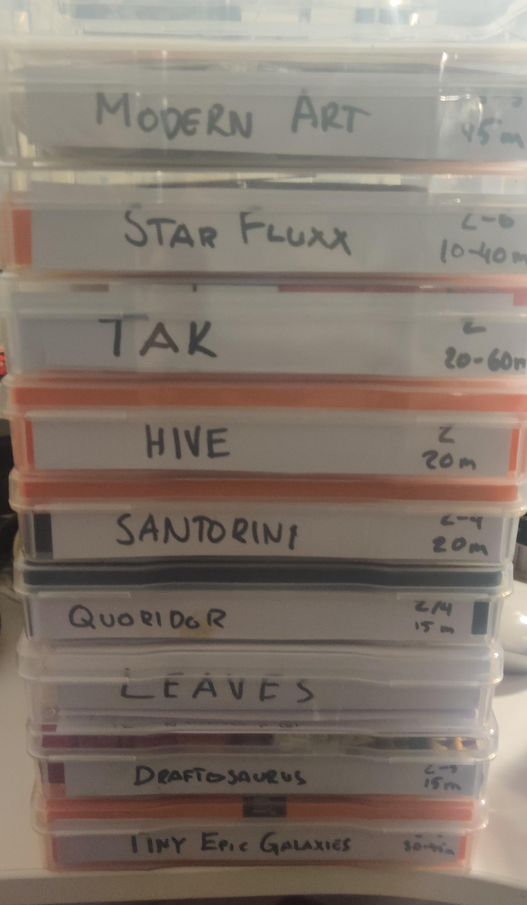 A stack of makeshift containers for board games in clear plastic photo containers with handwritten spines including game names for Modern Art, Star Fluxx, Tak, Hive, Santorini, Quoridor, Leaves, Draftosaurus and Tiny Epic Galaxies.