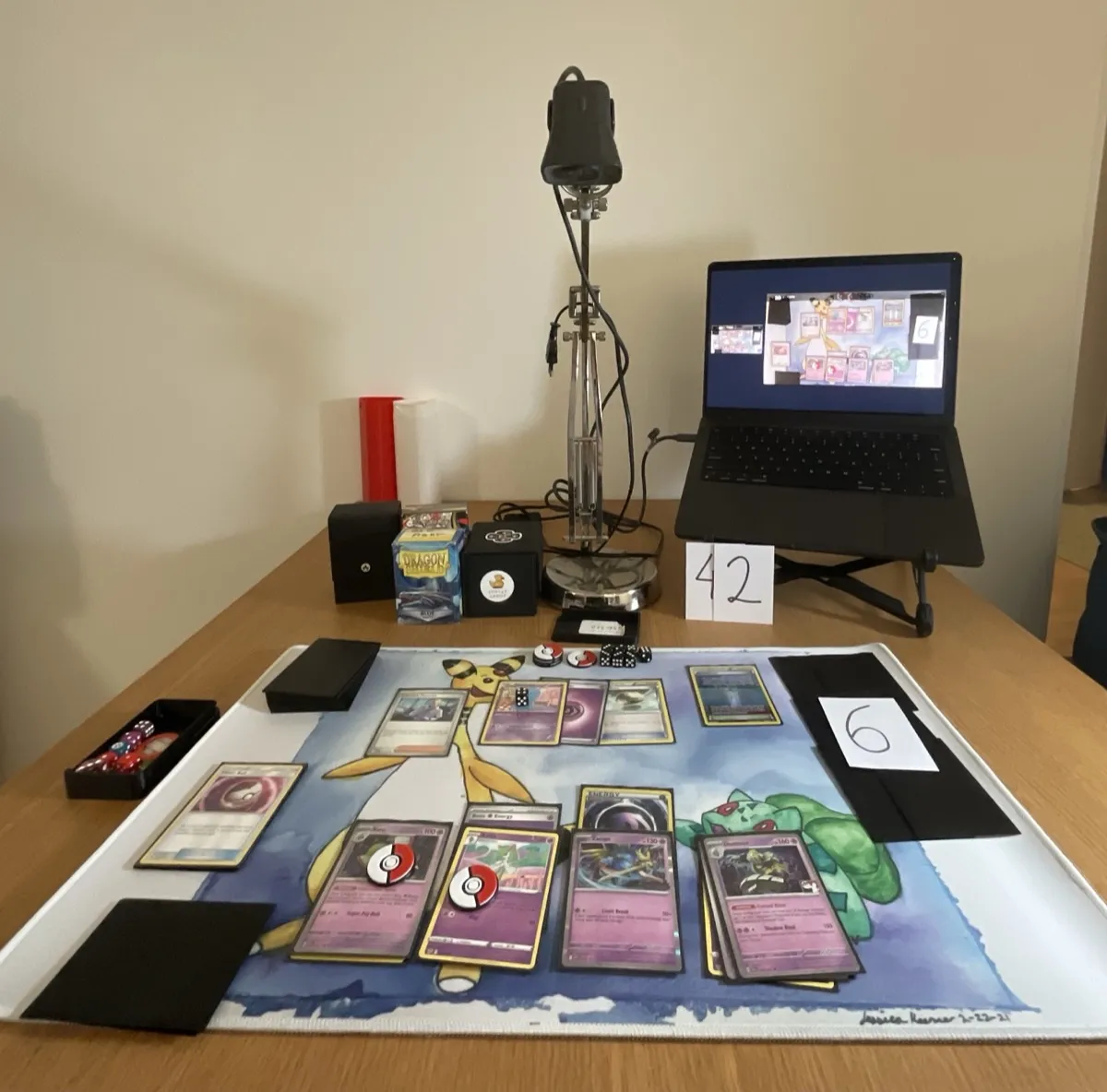 A desk with on-going Pokemon TCG game with a webcam pointing directly towards the playmat and a laptop open with SpellTable open and the webcam feed shown there.